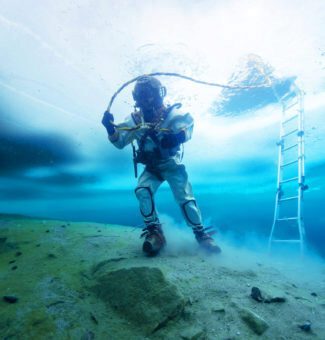 A diver in full gear sinks deeper beneath a frozen ice surface above.
