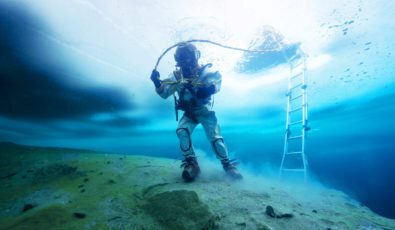 A diver in full gear sinks deeper beneath a frozen ice surface above.