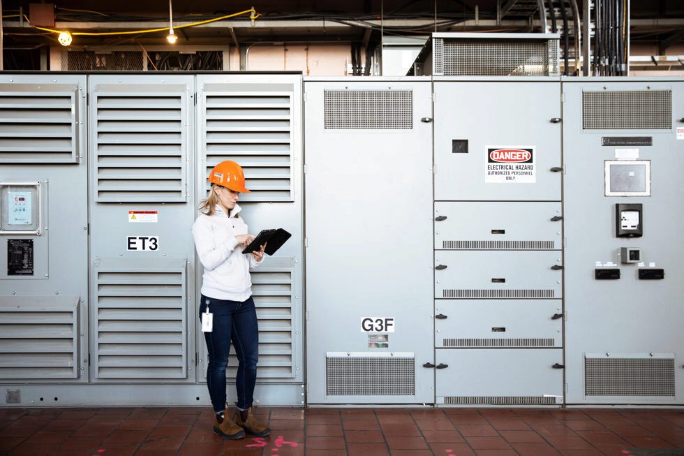 A female worker in a hard hat uses a tablet computer near a electrical components.