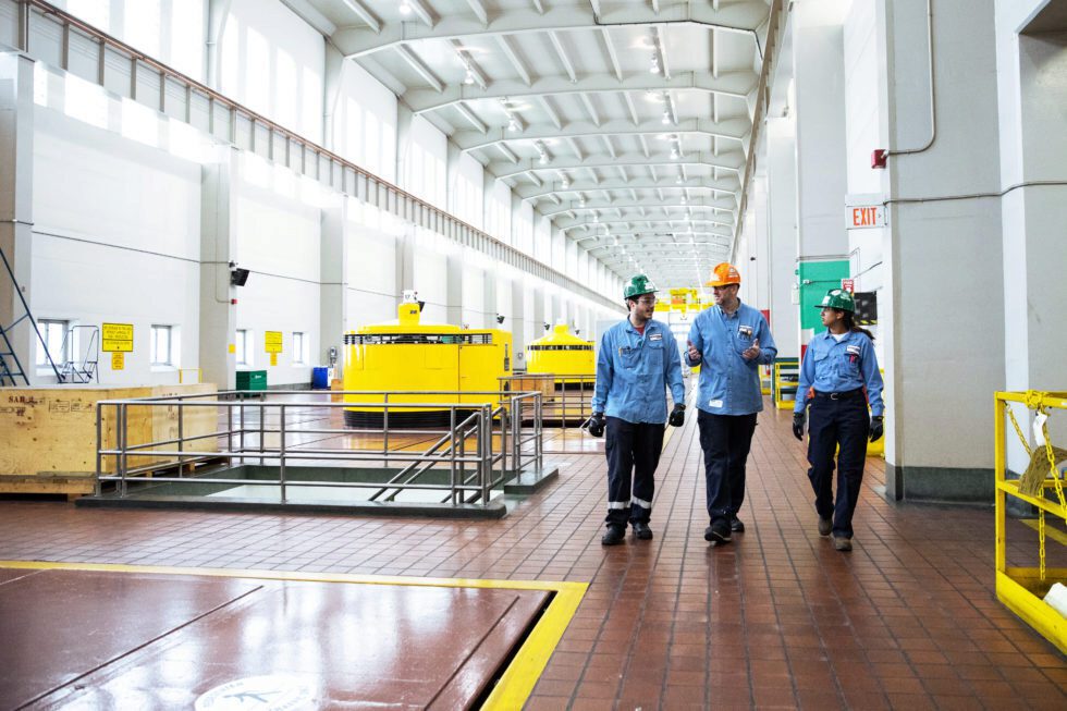 Three workers in hard hats talk while walking through a turbine hall.