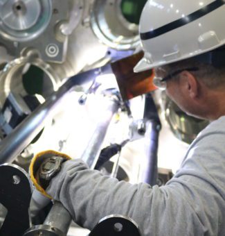 A male worker in a hard hat and safety glasses prepares to weld a nuclear unit bellows assembly.