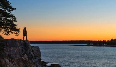 A hiker's sunset silhouetted at the edge of a lake shore.