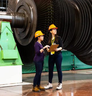 Two female workers in hard hats consult in front of a large nuclear turbine undergoing refurbishment.