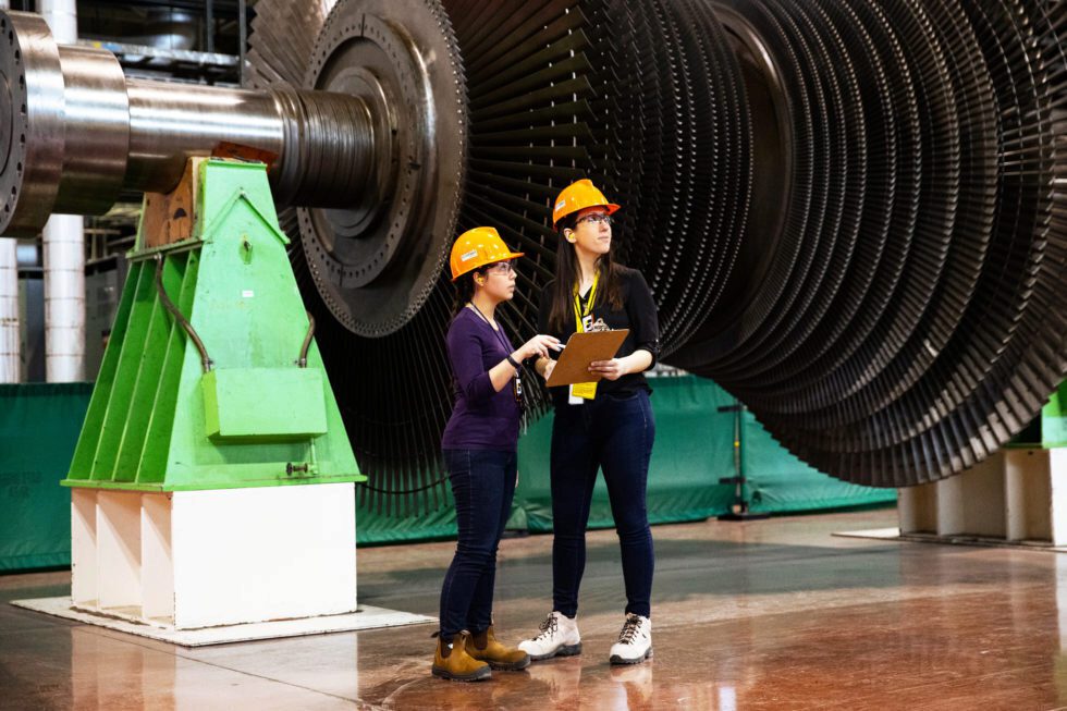 Two female workers in hard hats consult in front of a large nuclear turbine undergoing refurbishment.
