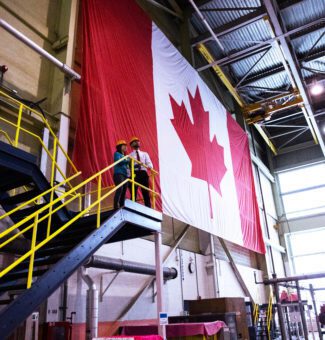 Workers in hard hats look out from the top of an open stairway before a huge Canadian flag in a nuclear plant.