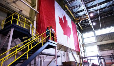 Workers in hard hats look out from the top of an open stairway before a huge Canadian flag in a nuclear plant.