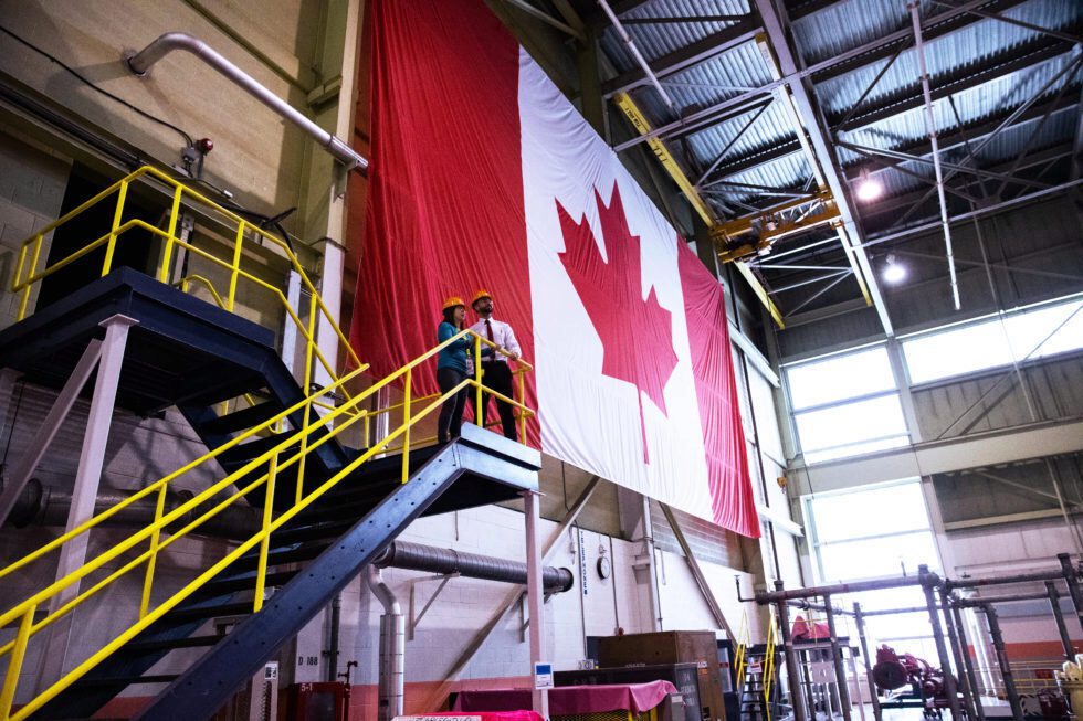 Workers in hard hats look out from the top of an open stairway before a huge Canadian flag in a nuclear plant.