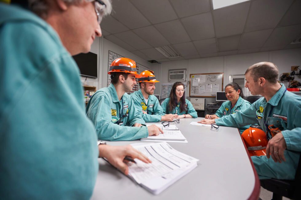 Employees in coveralls reviewing documents in meeting around a table.