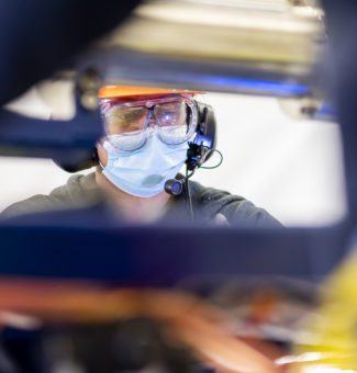 A worker in a mask and safety goggles wearing a telecommunications headset.