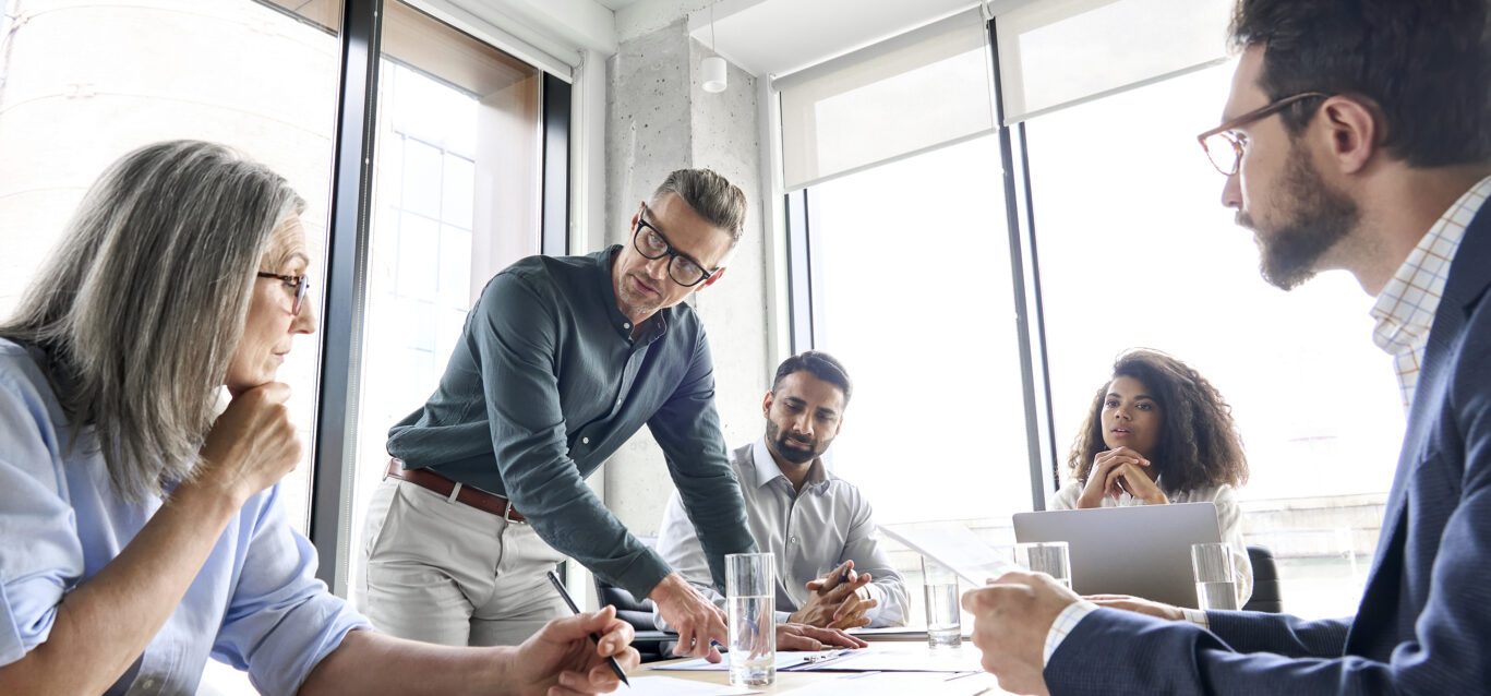 Male mature caucasian ceo businessman leader with diverse coworkers team, executive managers group at meeting. Multicultural professional businesspeople working together on research plan in boardroom.