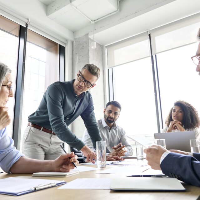 Male mature caucasian ceo businessman leader with diverse coworkers team, executive managers group at meeting. Multicultural professional businesspeople working together on research plan in boardroom.