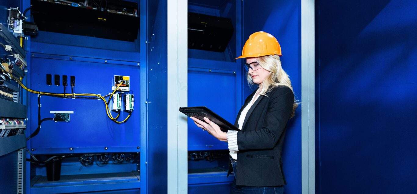A female worker in a hard hat uses a tablet computer near a electrical components.