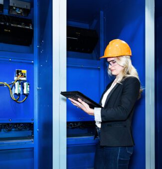 A female worker in a hard hat uses a tablet computer near a electrical components.