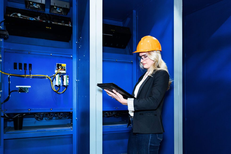 A female worker in a hard hat uses a tablet computer near a electrical components.