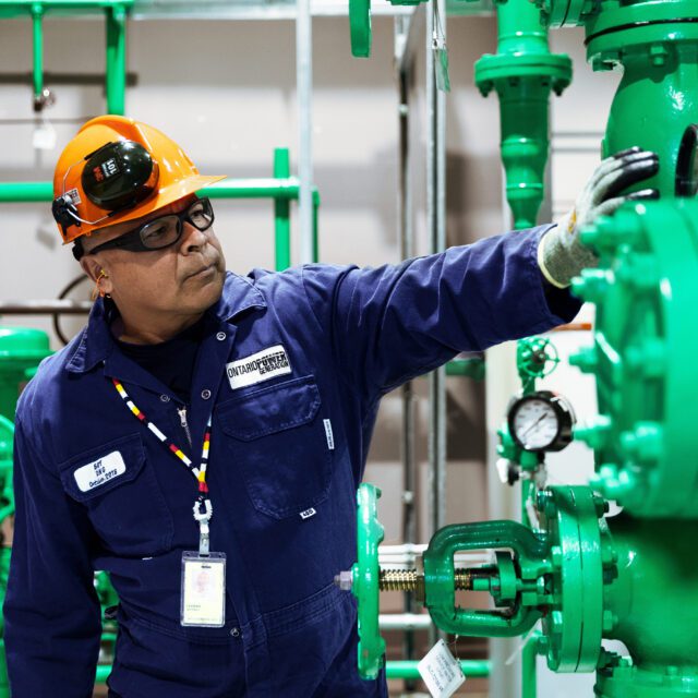 A male indigenous worker in coveralls and a hard hat examines valves in a power station.