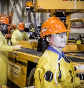 A female worker in protective plastic clothing, hart hat and safety glasses looking ahead with similar workers in the background.