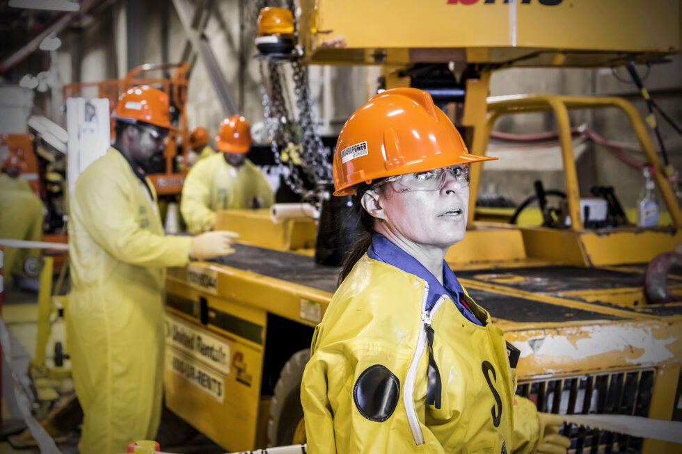A female worker in protective plastic clothing, hart hat and safety glasses looking ahead with similar workers in the background.