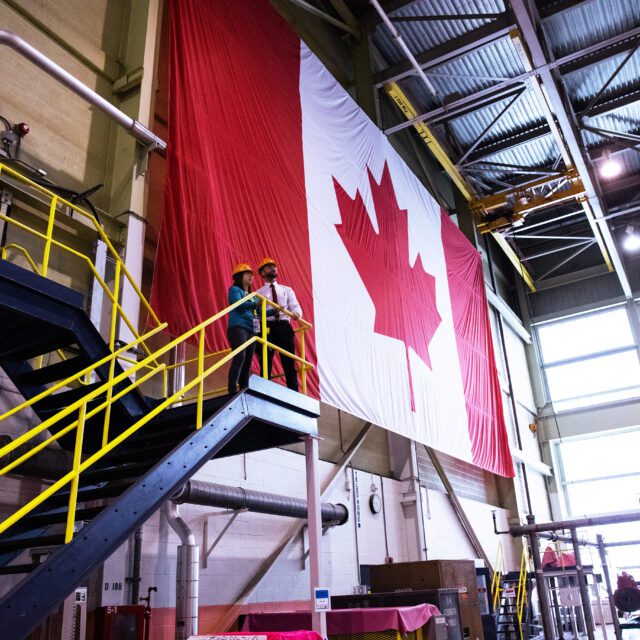 Workers in hard hats look out from the top of an open stairway before a huge Canadian flag in a nuclear plant.