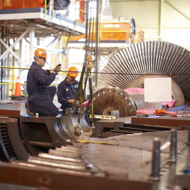 Two men in blue overalls and orange hard hats work on large machine parts.