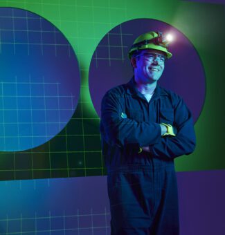 A worker in blue coveralls and a hard hat stands smiling with arms crossed.