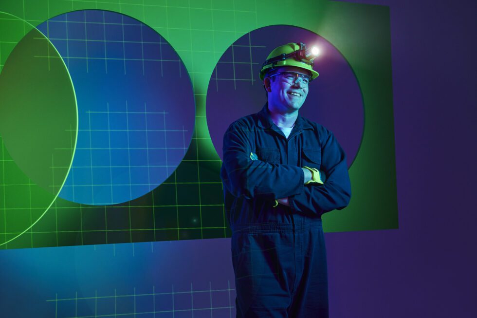 A worker in blue coveralls and a hard hat stands smiling with arms crossed.