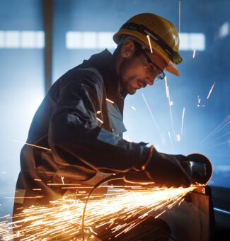 Heavy Industry Engineering Factory Interior with Industrial Worker Using Angle Grinder and Cutting a Metal Tube. Contractor in Safety Uniform and Hard Hat Manufacturing Metal Structures.