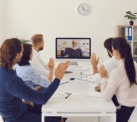 A group of people sit at a table interacting with person displayed on a computer screen.