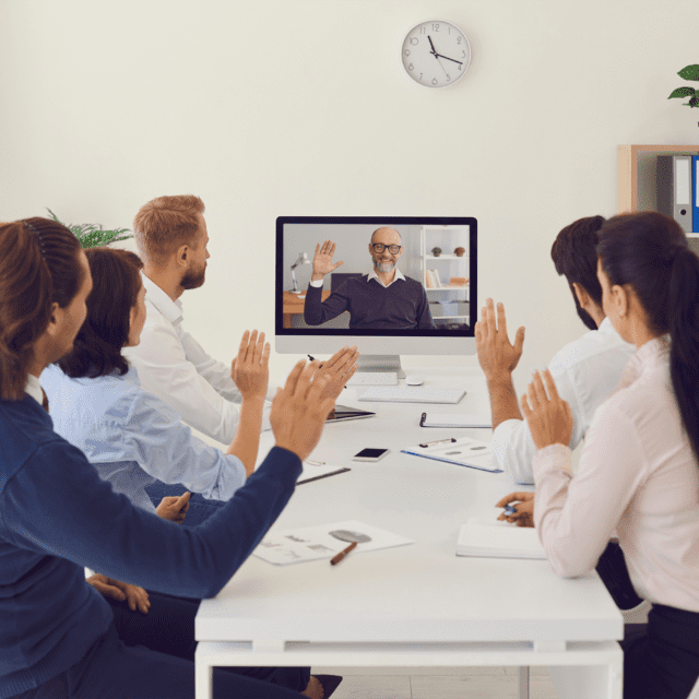 A group of people sit at a table interacting with person displayed on a computer screen.