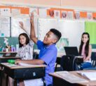 A child at a desk in a classroom raises his hand to ask a question.