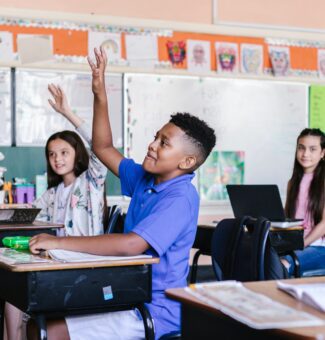 A child at a desk in a classroom raises his hand to ask a question.