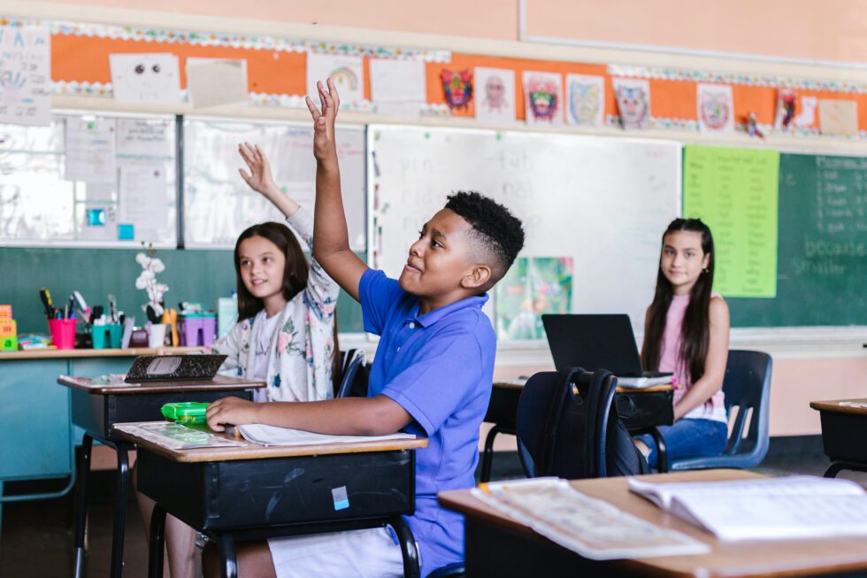 A child at a desk in a classroom raises his hand to ask a question.