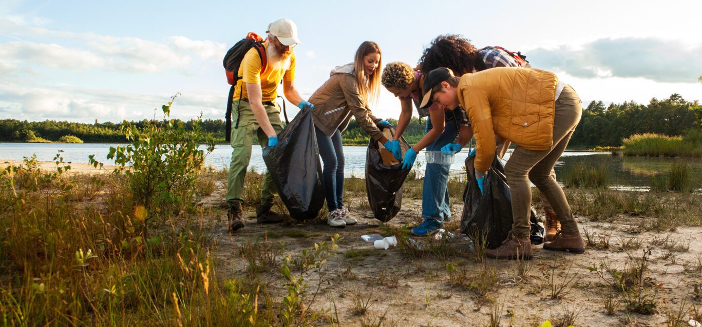 Young multi ethnic group of volunteers cleaning up the shore of a forest lake together, they are collecting trash and holding garbage bags. Concept of clean nature, ecology and sustainability.
