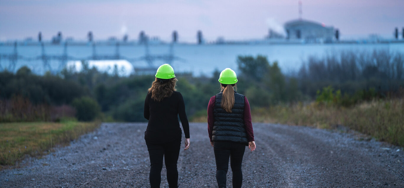 Two employees in hard hats walk on a gravel road outside near the site of the Darlington New Nuclear Project.