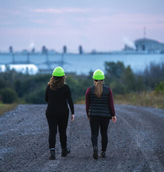 Two employees in hard hats walk on a gravel road outside near the site of the Darlington New Nuclear Project.