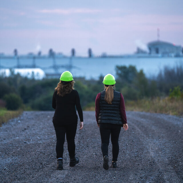 Two employees in hard hats walk on a gravel road outside near the site of the Darlington New Nuclear Project.