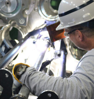 A male worker in a hard hat and safety glasses prepares to weld a nuclear unit bellows assembly.